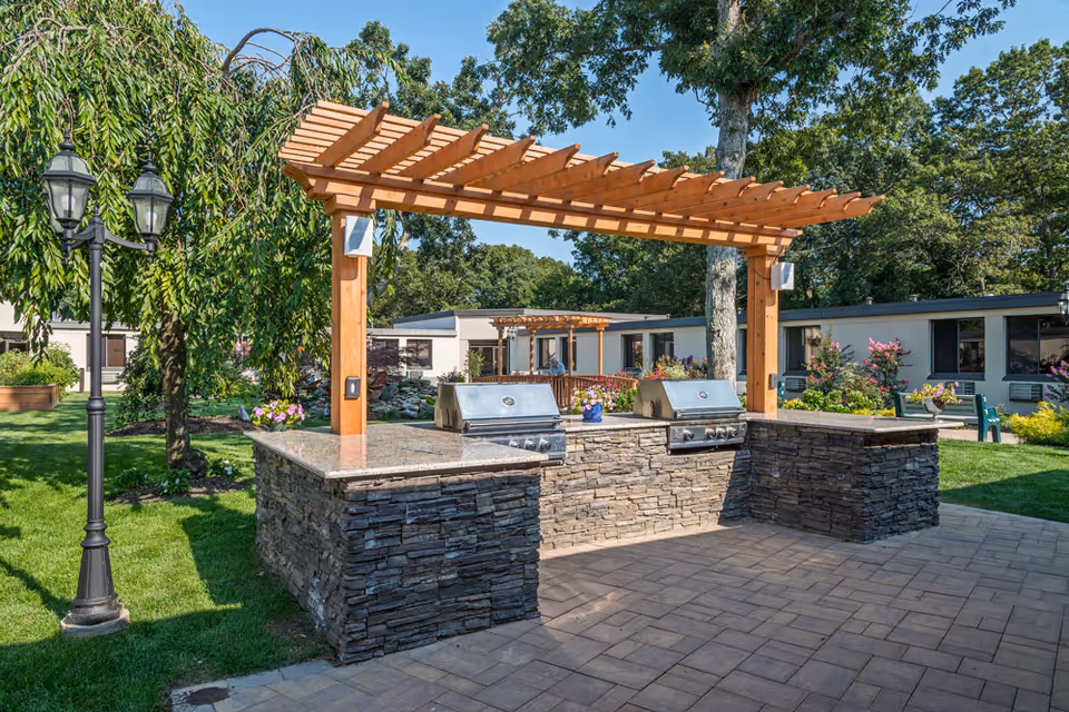 Outdoor barbecue area with two stainless steel grills set in a stone countertop under a wooden pergola. The area is surrounded by green grass, trees, and shrubs with a clear blue sky overhead. A black vintage-style street lamp is visible on the left side.