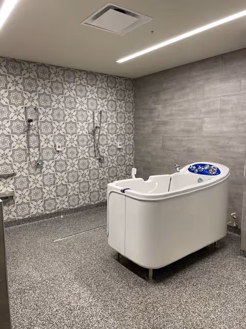 A therapy bathing room with a white walk-in bathtub, patterned tile wall with multiple showerheads, and gray flooring.