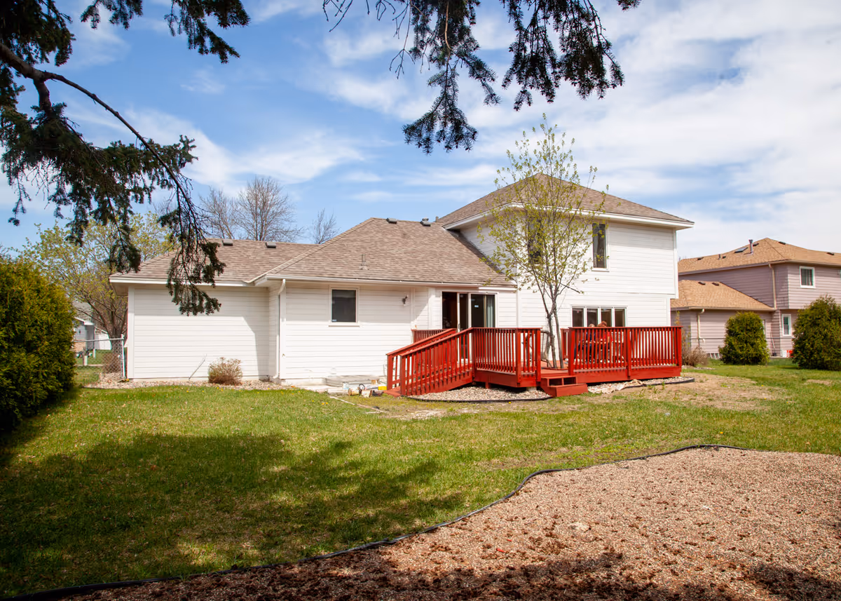 View of a white two-story house with a red wooden deck and a grassy backyard under a blue sky.