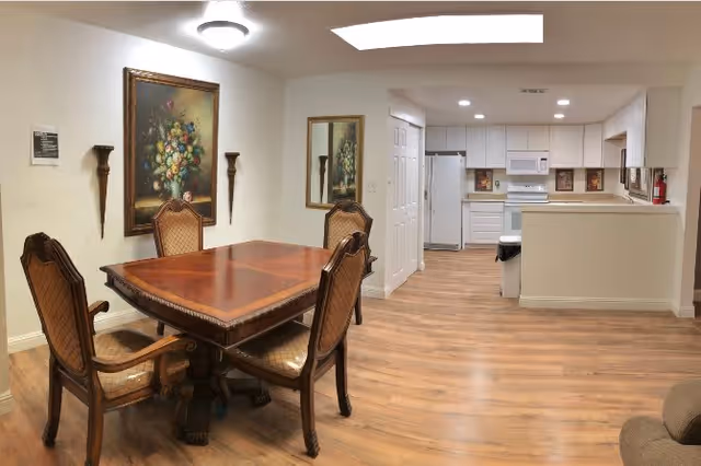 A dining area with a wooden table and four upholstered chairs. The walls are decorated with a large floral painting and a mirror. The room has wooden flooring and leads into a kitchen with white cabinets, a refrigerator, microwave, stove, and a trash can. The ceiling has recessed lighting and a skylight.