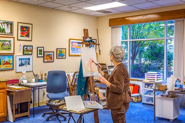 An elderly woman painting at an easel in a bright art room with framed artwork on the walls, supply tables, and a large window overlooking greenery.