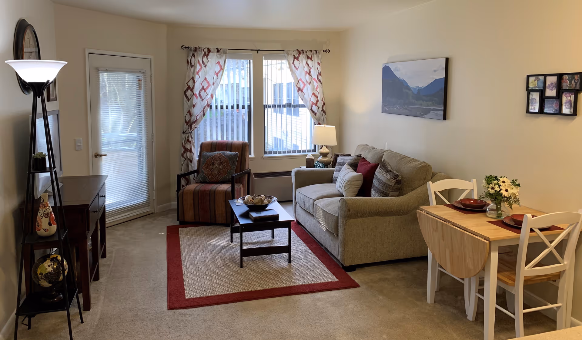 A cozy living room area with a beige sofa adorned with multiple cushions, a striped armchair, a small coffee table on a beige and red rug, and a floor lamp. There is a small dining table with two chairs set with red placemats and a vase of flowers. The room has a window with patterned curtains and a glass door with blinds. Wall decorations include a landscape painting and a photo collage frame.