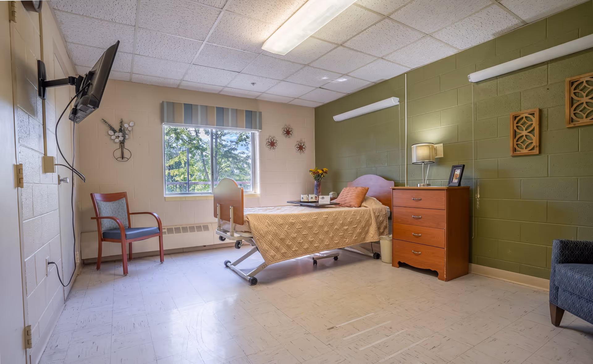 A simple, clean bedroom in a healthcare facility with a single bed covered in a beige quilt, a wooden dresser with a lamp and framed photo, a window with a view of trees, a mounted TV on the wall, and two chairs placed on opposite sides of the room. The walls are painted beige and green with decorative wall hangings.