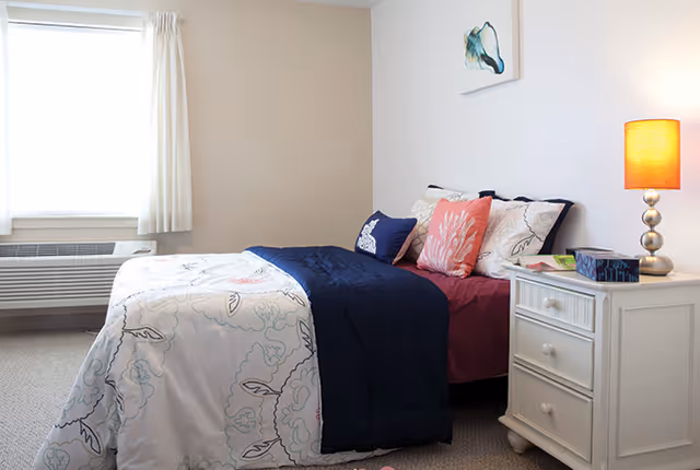 A neatly made single bed with floral-patterned bedding, a navy throw and decorative pillows next to a white nightstand with a lamp and a window.