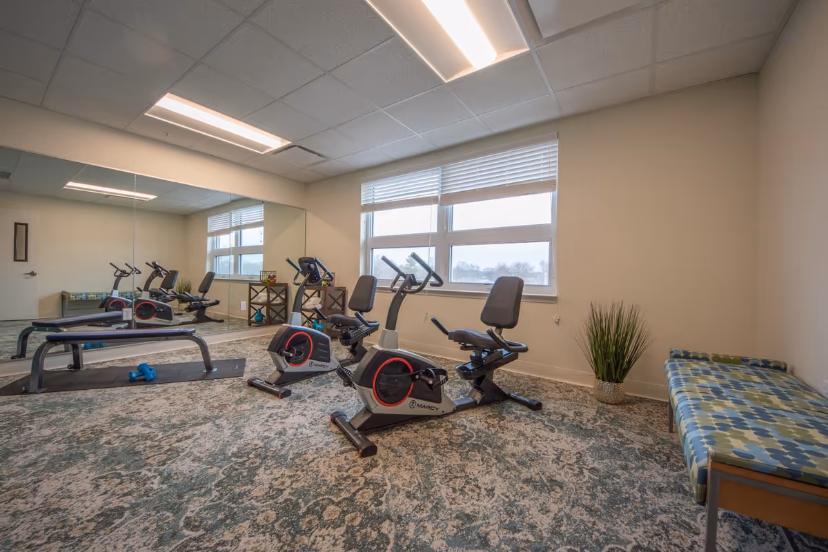 A small exercise room with two recumbent stationary bikes, a workout bench, large wall mirror, and a cushioned bench by a window.
