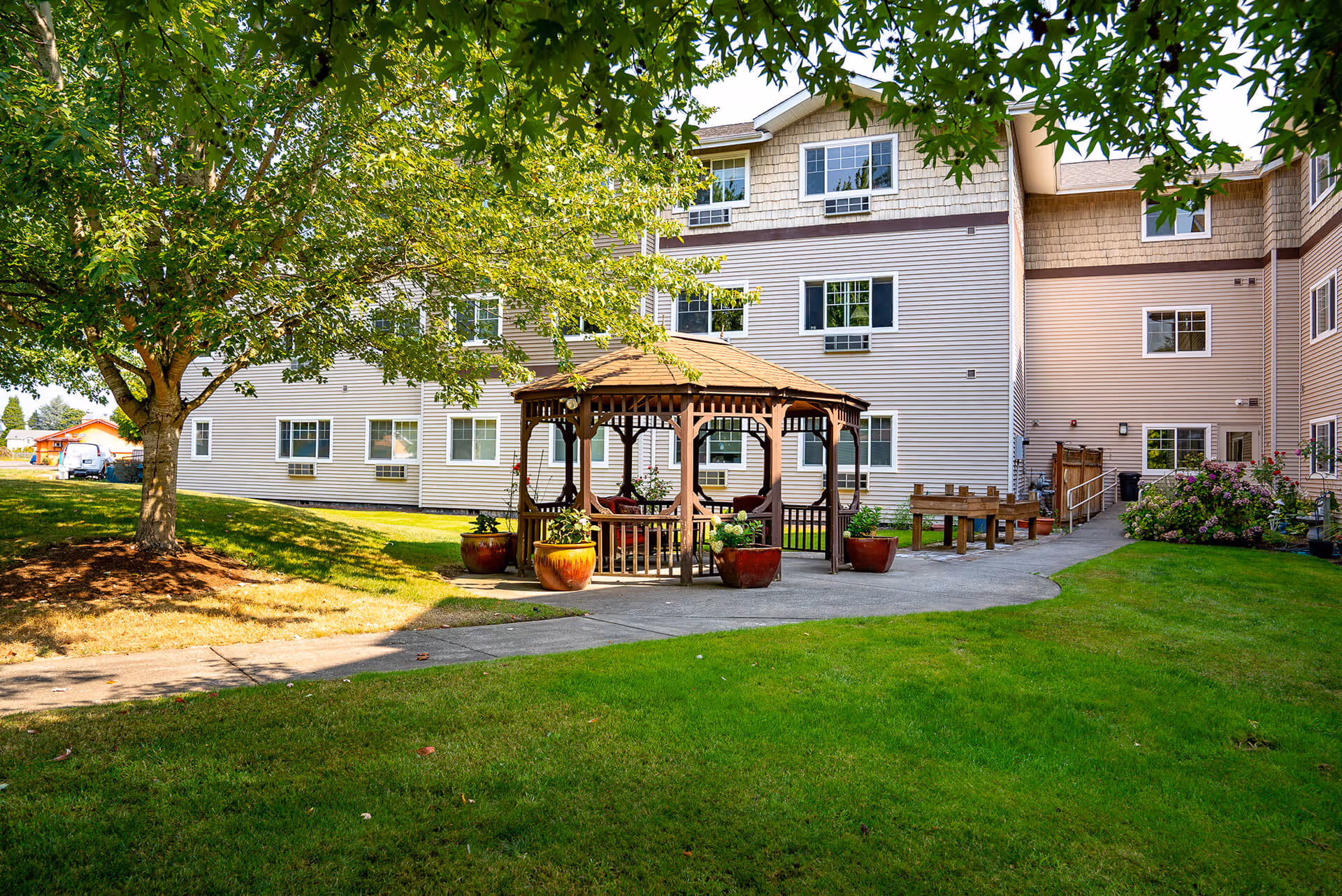 Three-story senior living building courtyard with a wooden gazebo, potted plants, benches, and a grassy lawn under trees.