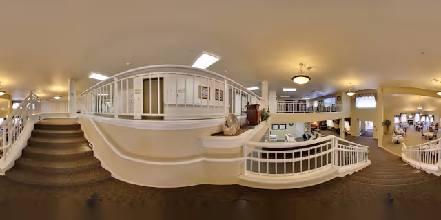 Panoramic interior of a senior living facility lobby with carpeted staircases, a reception desk, and seating areas across two levels.