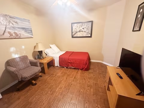 A small bedroom with a single bed covered with a red blanket and white pillows against a beige wall. Next to the bed is a wooden nightstand with a lamp and a small decorative item. A patterned armchair is positioned near the nightstand. On the opposite side of the room, there is a wooden TV stand with a flat-screen television and a remote control. The room has wooden flooring and two framed pictures hanging on the walls.