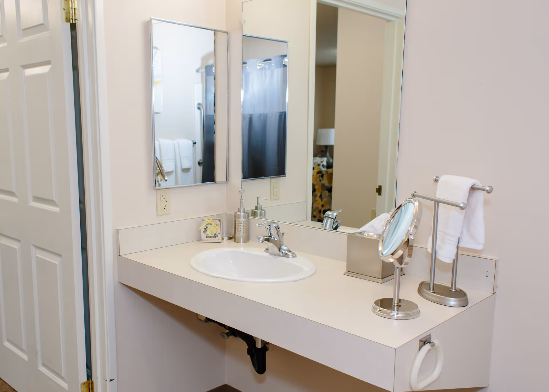 A clean bathroom vanity with a white sink, a silver faucet, a small round makeup mirror, a tissue box, a soap dispenser, and a towel holder with a white towel. There are two rectangular mirrors on the wall above the sink, and a partially open door to the left reveals a shower curtain and towels in the adjacent bathroom area.