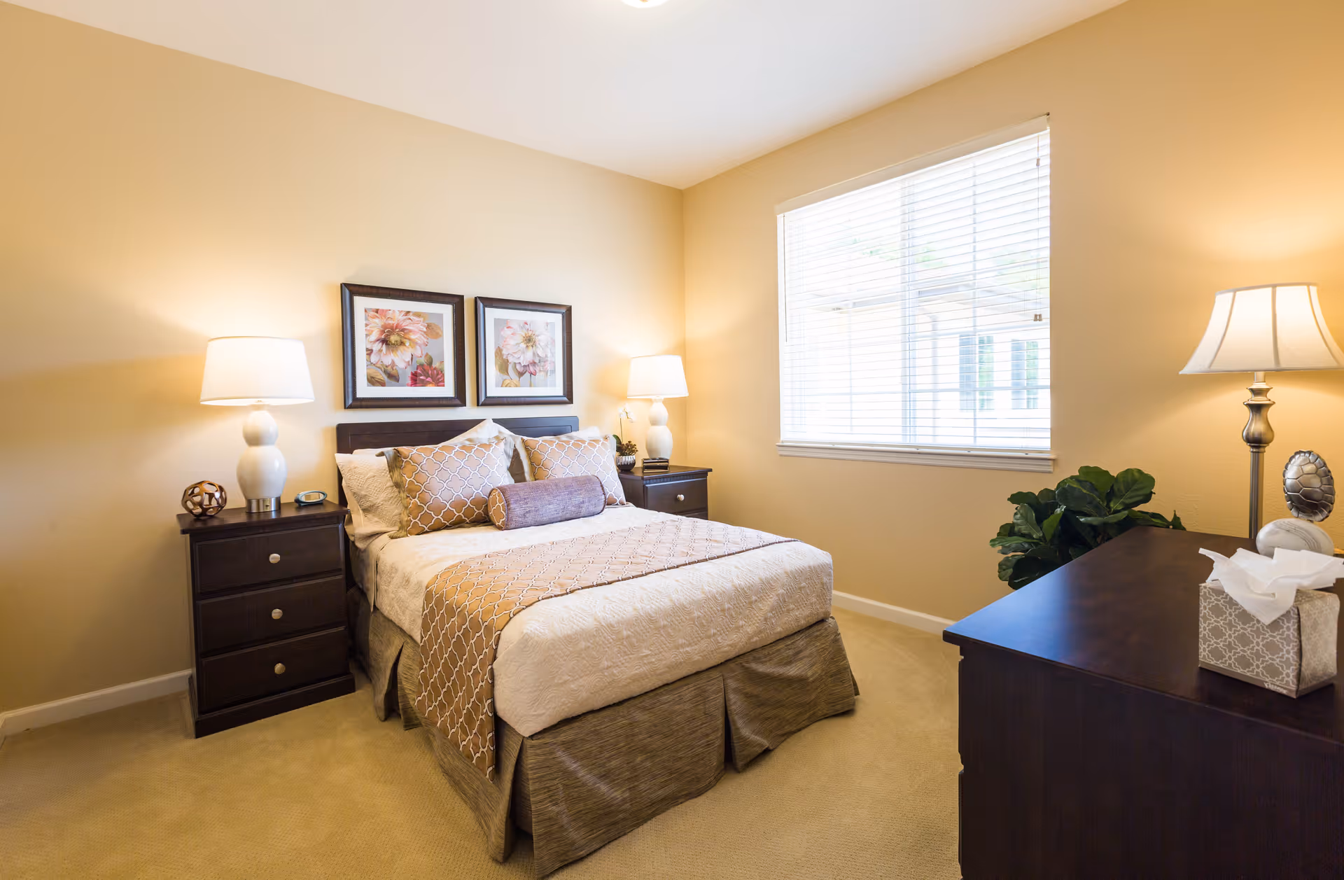 A neatly made bed with beige and brown bedding in a well-lit bedroom. The room features two dark wooden nightstands with white lamps on each side of the bed, two framed floral artworks above the headboard, a window with white blinds, a dark wooden dresser with a lamp, a tissue box, and a decorative plant in the corner.
