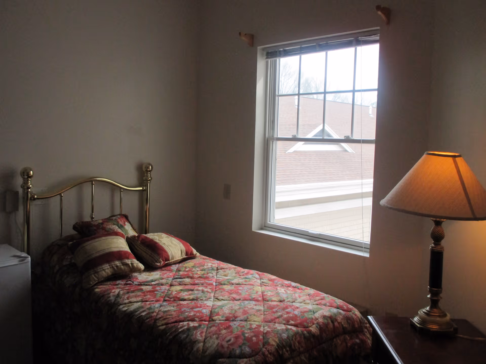 A small bedroom with a single bed featuring a floral patterned bedspread and three pillows. The bed has a brass headboard. Next to the bed is a wooden nightstand with a lamp that has a beige lampshade. There is a window with white trim letting in natural light, showing part of a roof and some trees outside. The walls are plain and light-colored.