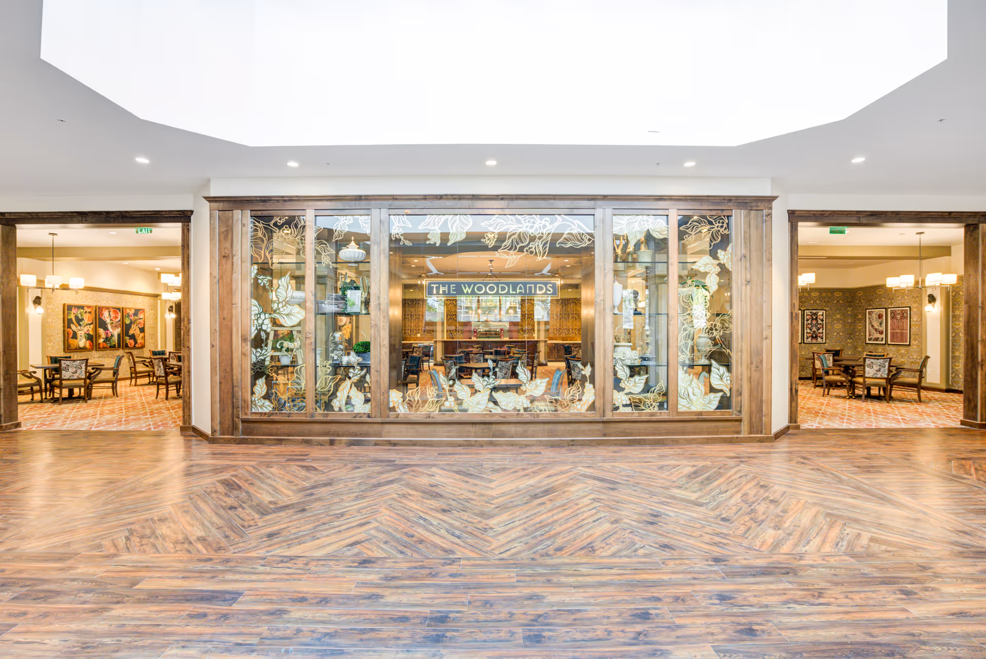 Interior view of a senior living facility hallway with wooden flooring and a large glass display case in the center featuring floral designs and the sign 'The Woodlands'. On either side of the display case are dining areas with tables, chairs, and decorative wall art.