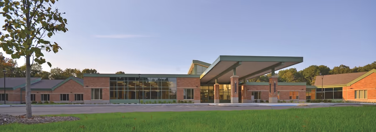 Low-rise brick building front with large glass windows and a covered porte-cochère entrance set behind a grassy lawn.