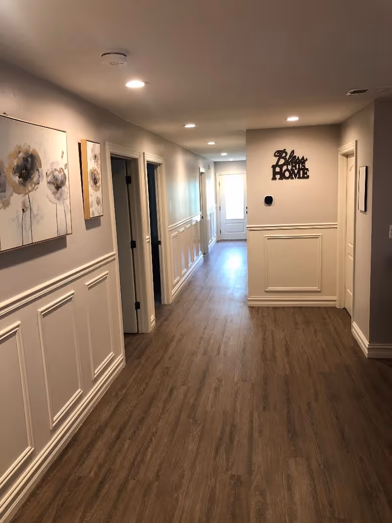 Long, well-lit interior hallway with wood-look flooring, white wainscoting, wall art, and several doorways.