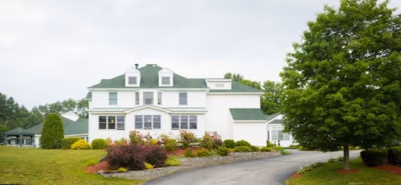 White multi-story nursing home with green roof, landscaped grounds, and surrounding trees.