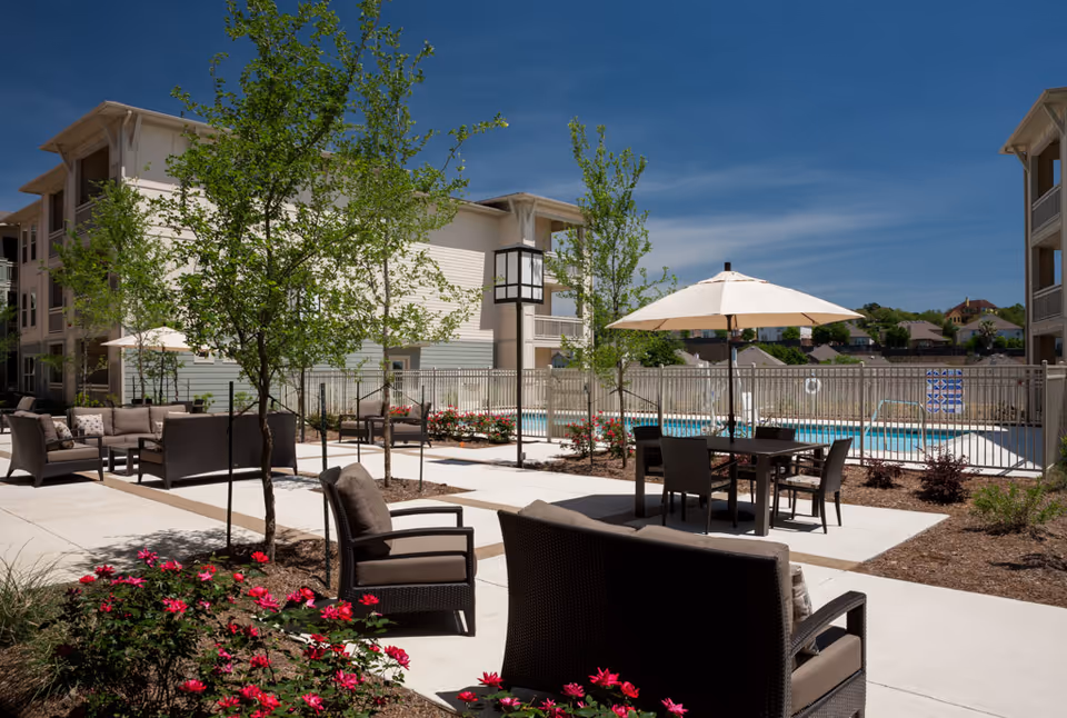 Outdoor courtyard of a senior living community with patio seating, umbrellas, landscaping, and a fenced swimming pool between residential buildings.