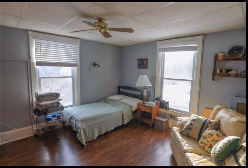 A cozy bedroom with two large windows letting in natural light. The room features a single bed with light green bedding, a wooden nightstand with a lamp and some items on it, a beige leather couch with multiple decorative pillows, a ceiling fan with wooden blades, and a small shelving unit with folded blankets and towels. The walls are painted light gray and the floor is wooden.