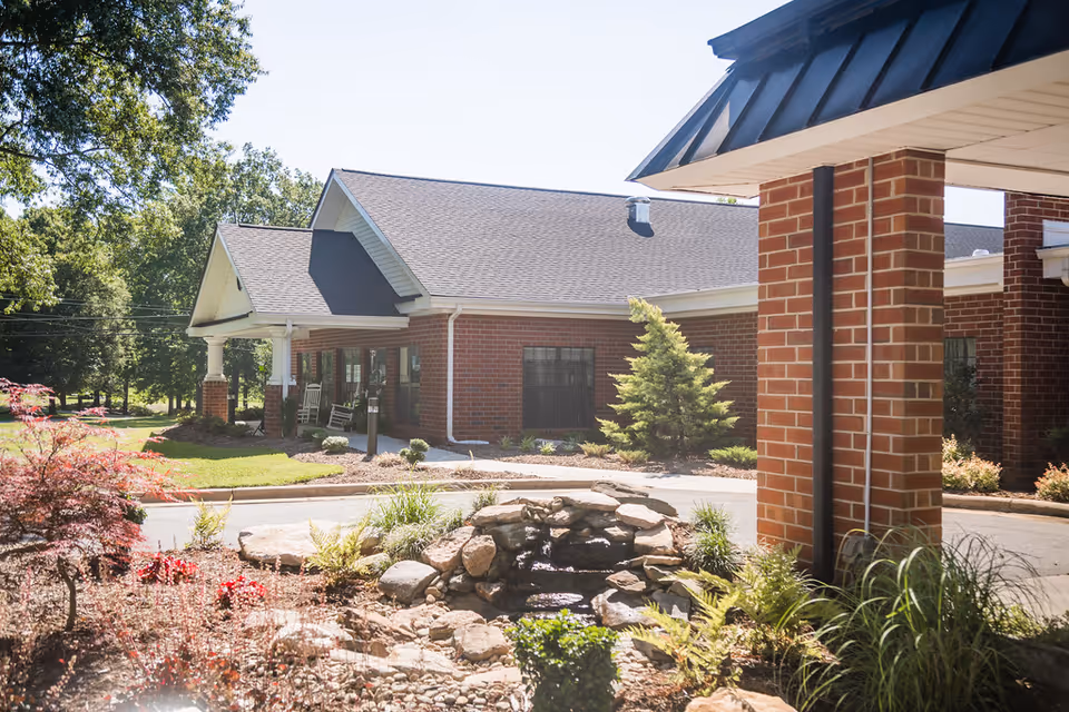 Exterior view of a brick building with a covered entrance supported by brick columns. In front of the building, there is a landscaped garden area with rocks, plants, and a small water feature. Trees and greenery surround the building under a clear sky.