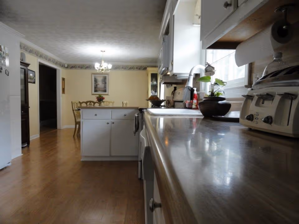 View of a kitchen with white cabinets and a countertop on the right side, a sink under a window with a plant on the counter, and a dining area with a table and chairs in the background under a chandelier light fixture.