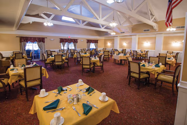 Spacious dining room with multiple tables set with yellow tablecloths, green napkins, and chairs under exposed white ceiling beams.