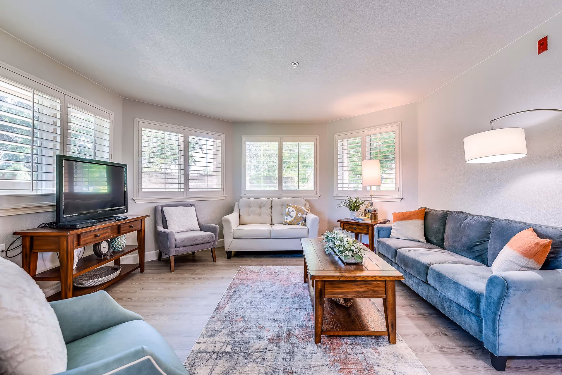 Bright furnished living room with a sofa, loveseat, armchairs, wooden coffee table, TV on a console, and large shuttered windows.