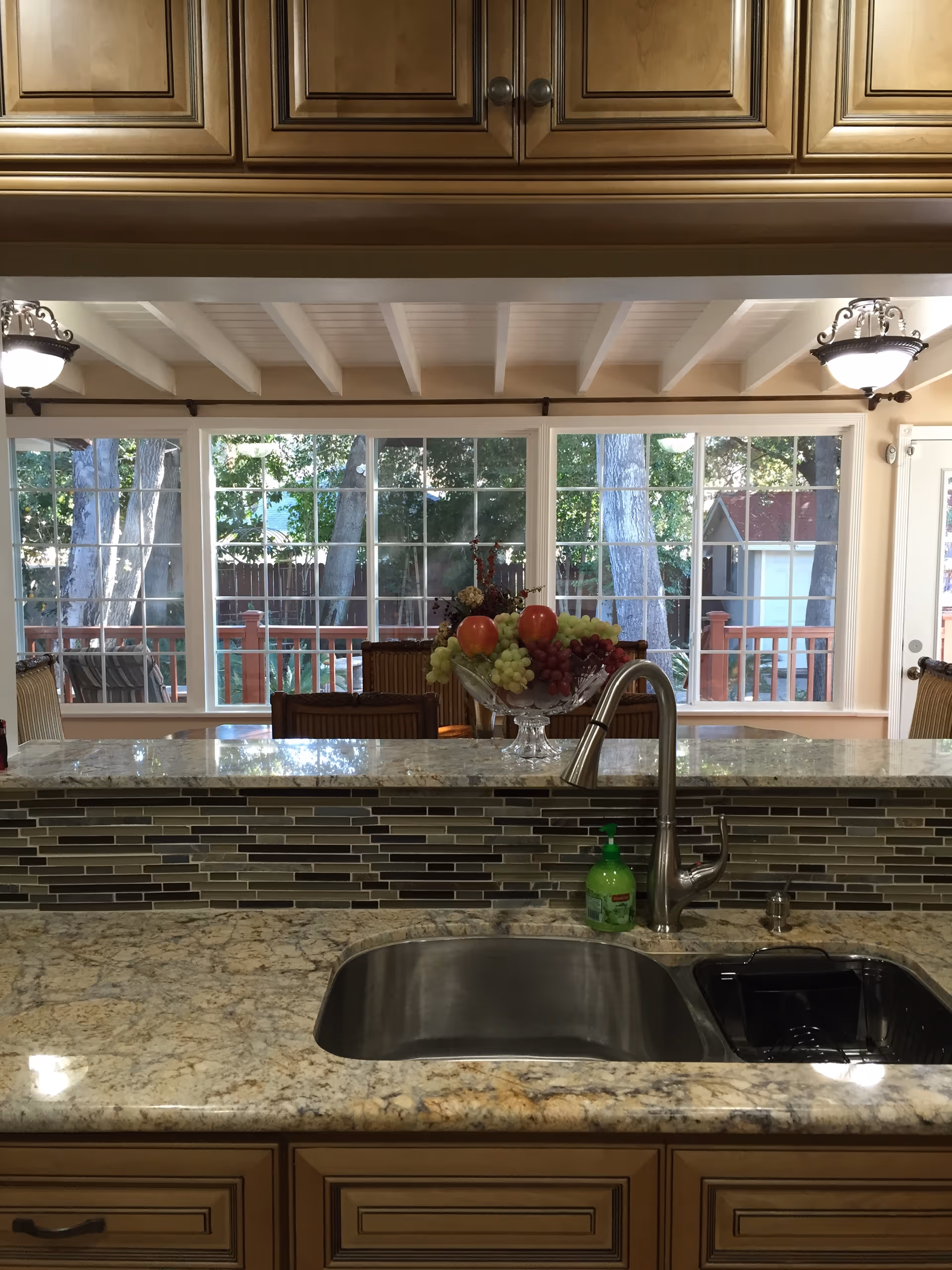 View of a kitchen sink with a modern faucet set in a granite countertop. Behind the sink is a raised counter with a decorative glass bowl filled with assorted fruits. Large windows in the background reveal a wooden deck and trees outside, allowing natural light to fill the space. The ceiling features exposed beams and two hanging light fixtures.