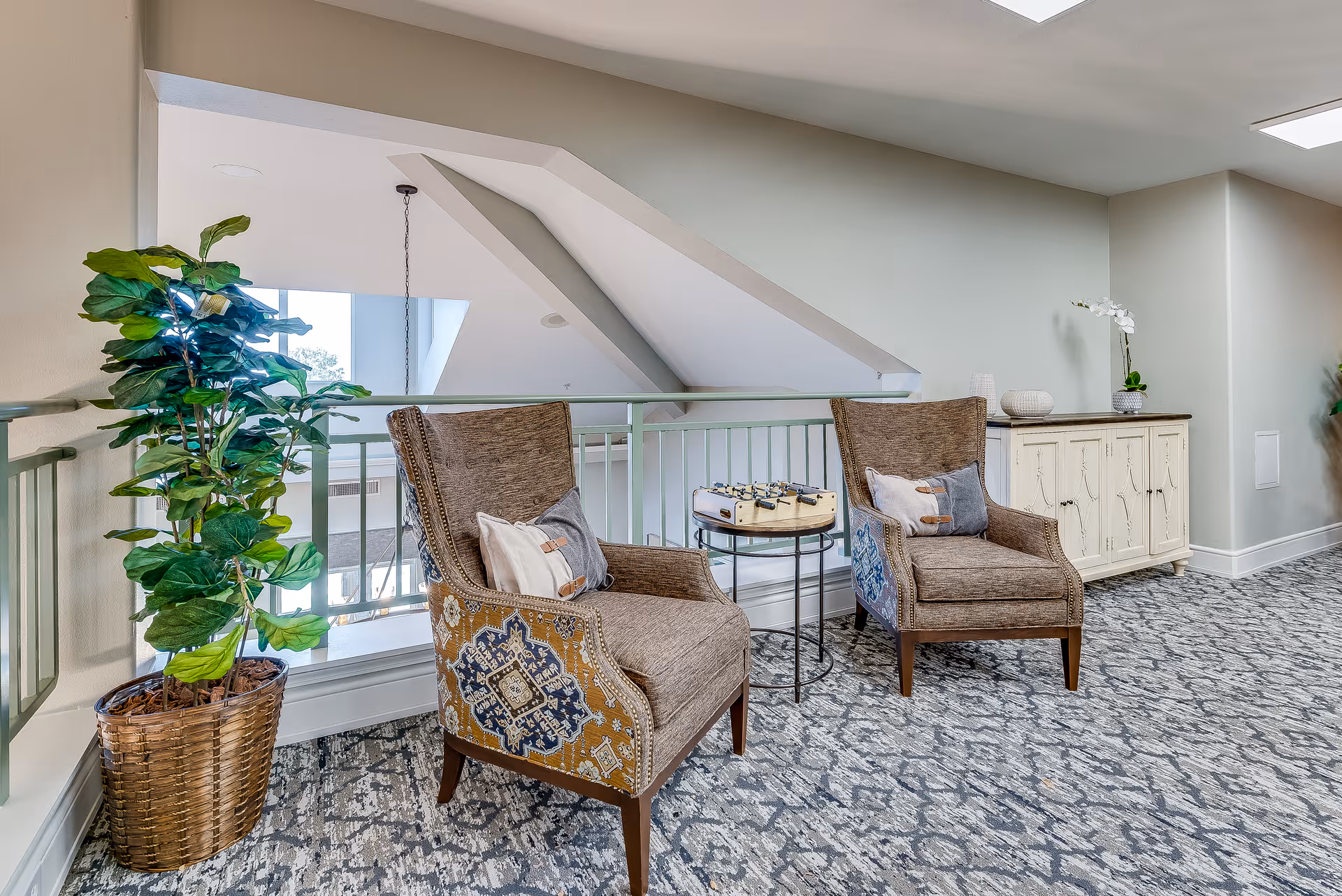 A cozy seating area in a senior living facility featuring two upholstered armchairs with patterned sides and decorative pillows, a small round table with a foosball game on top, a large potted plant, and a white sideboard with decorative items against a light-colored wall. The area has patterned carpet flooring and a railing overlooking a lower level with large windows.