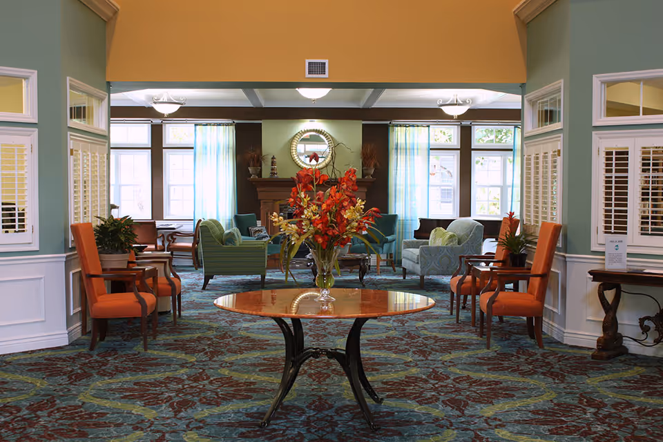 A spacious and well-lit living room area in an assisted living facility featuring a round wooden table with a vase of red and yellow flowers in the center. The room has patterned carpet flooring, multiple upholstered chairs in orange and blue-green tones, large windows with sheer blue curtains, and a fireplace with a round mirror above it.