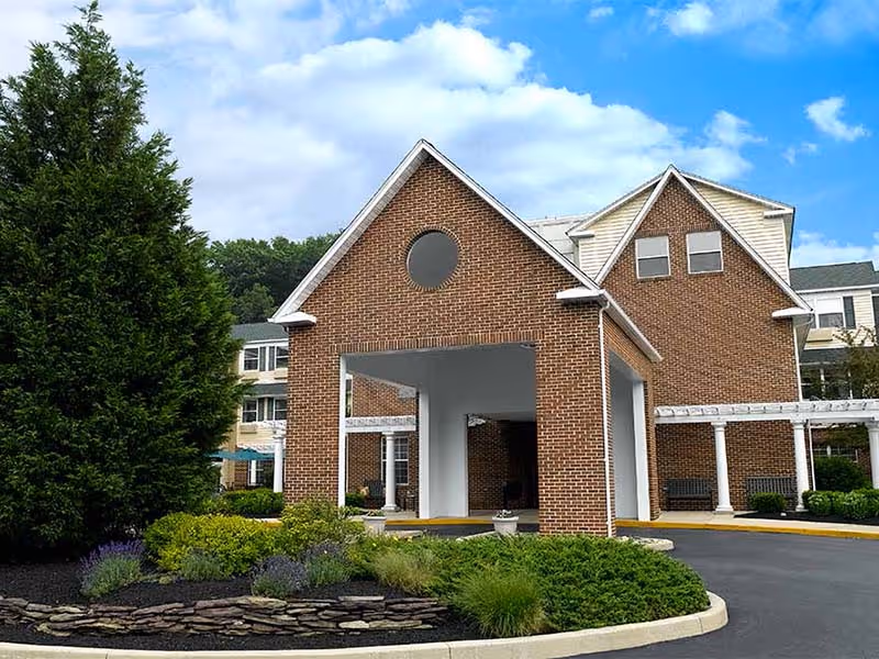 Exterior view of a senior living facility building with a brick facade, a covered entrance, and a landscaped area with bushes and a large tree under a partly cloudy sky.