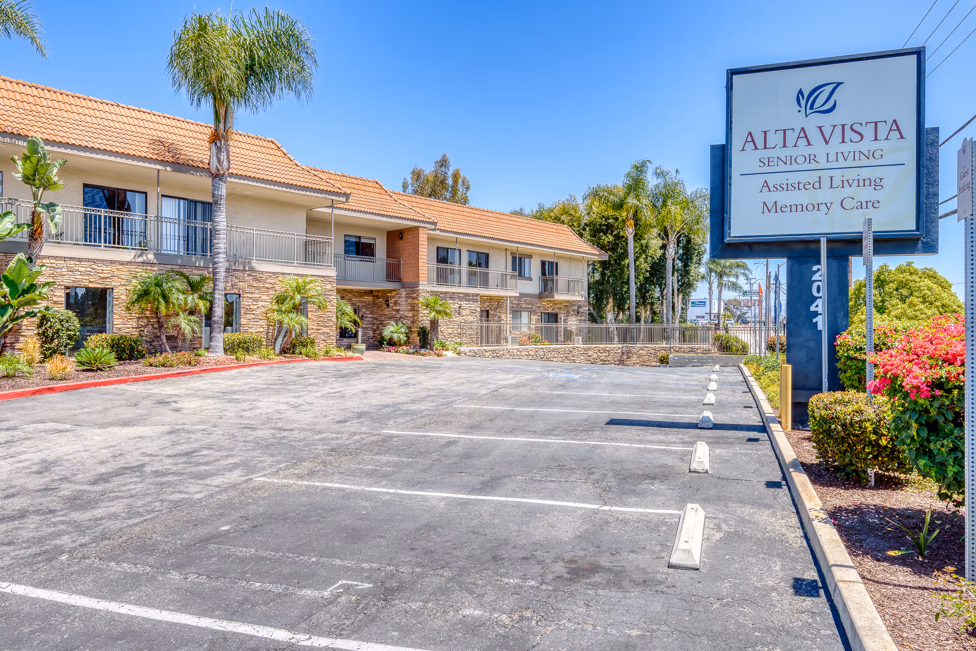 Exterior view of Alta Vista Senior Living facility with a two-story building featuring a tiled roof, stone facade, and balconies. There are palm trees and landscaped bushes around the building and an empty parking lot in the foreground. A large sign near the parking lot reads 'Alta Vista Senior Living Assisted Living Memory Care.'
