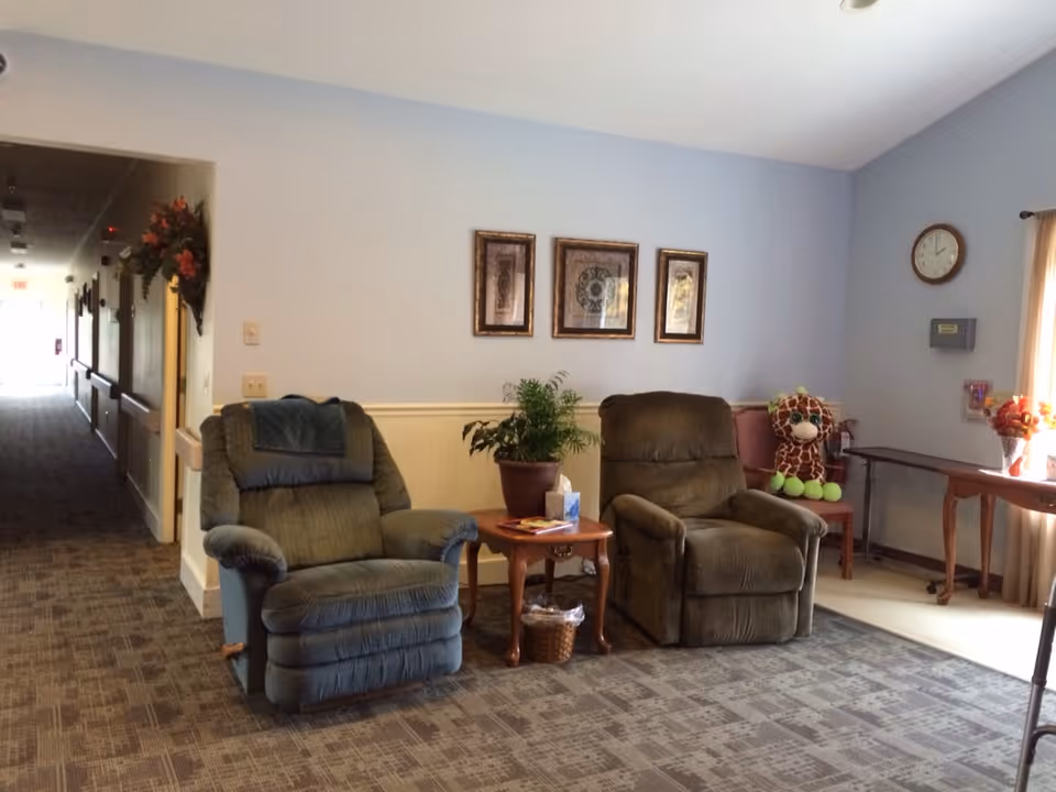 A cozy seating area in a senior living facility with two recliner chairs separated by a small wooden table holding a potted plant and tissues. Behind the chairs, three framed pictures hang on a light blue wall. To the right, a small table with a vase of flowers and a large stuffed giraffe toy sits near a window with beige curtains. A clock and a wall-mounted box are also visible on the wall. A hallway extends to the left with a floral wall decoration near the entrance.