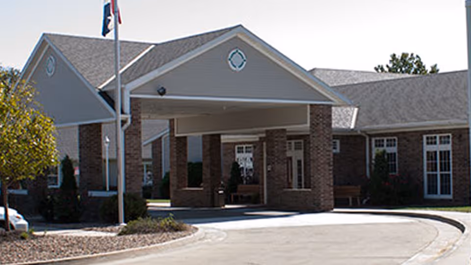 Exterior view of the entrance to Country Club Rehab & Healthcare Center, featuring a covered drop-off area supported by brick columns, a flagpole with a flag, and surrounding landscaping with trees and bushes.