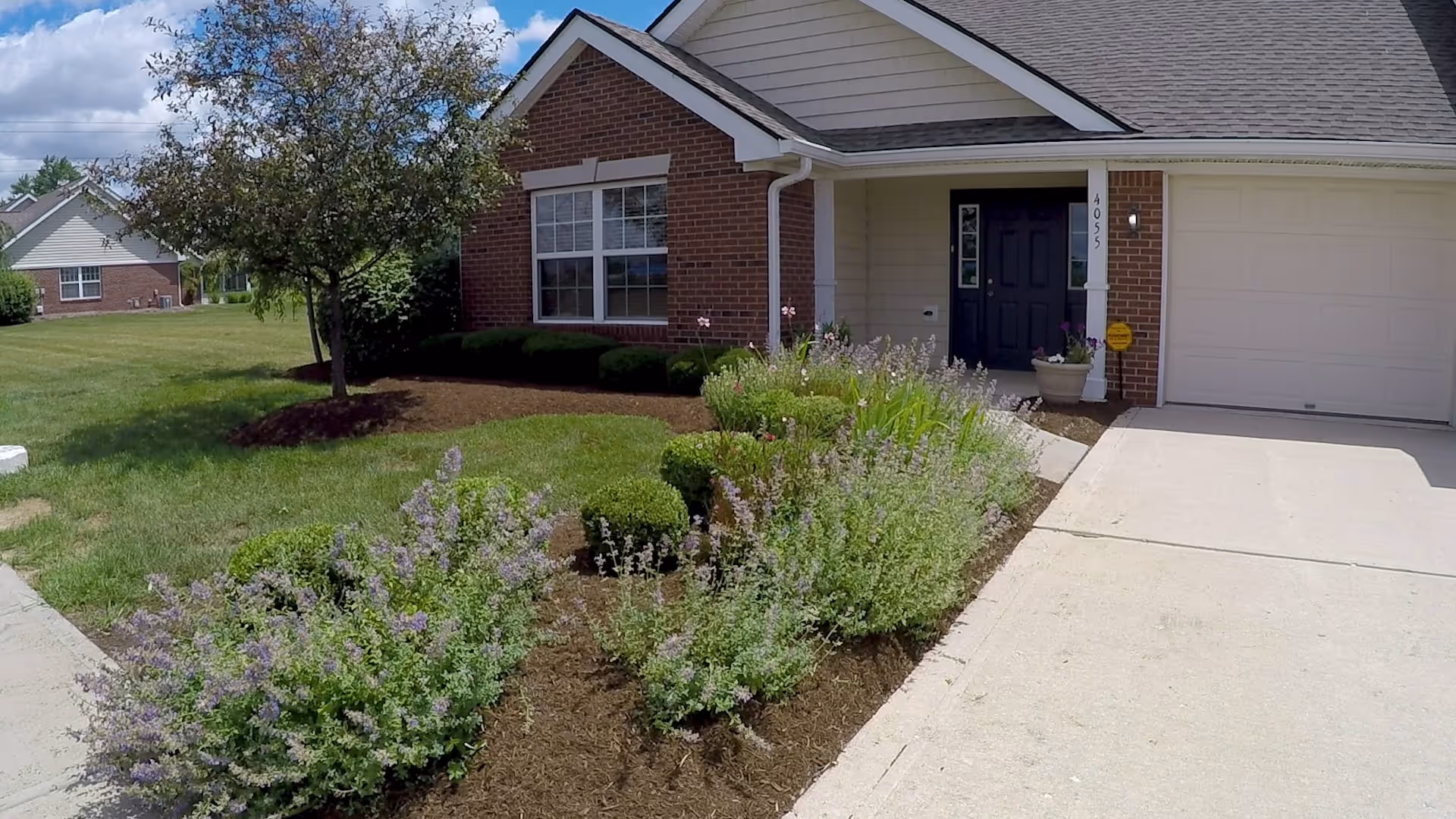 Front exterior view of a single-story brick house with a black front door, a garage, a tree, and a well-maintained garden with shrubs and flowering plants under a partly cloudy sky.