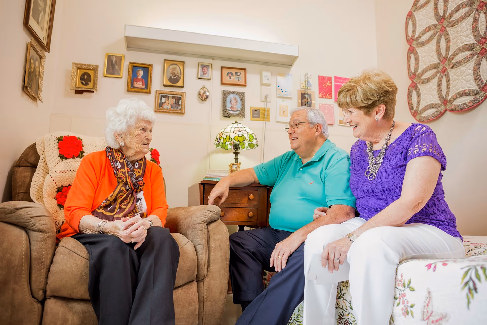 Three elderly people sitting and chatting in a cozy living room. An elderly woman with white hair and an orange cardigan sits in a recliner chair, while an elderly man in a turquoise polo shirt and an elderly woman in a purple top sit on a floral-patterned couch. The room has framed photos on the wall, a small table with a decorative lamp, and a quilt hanging on the wall.