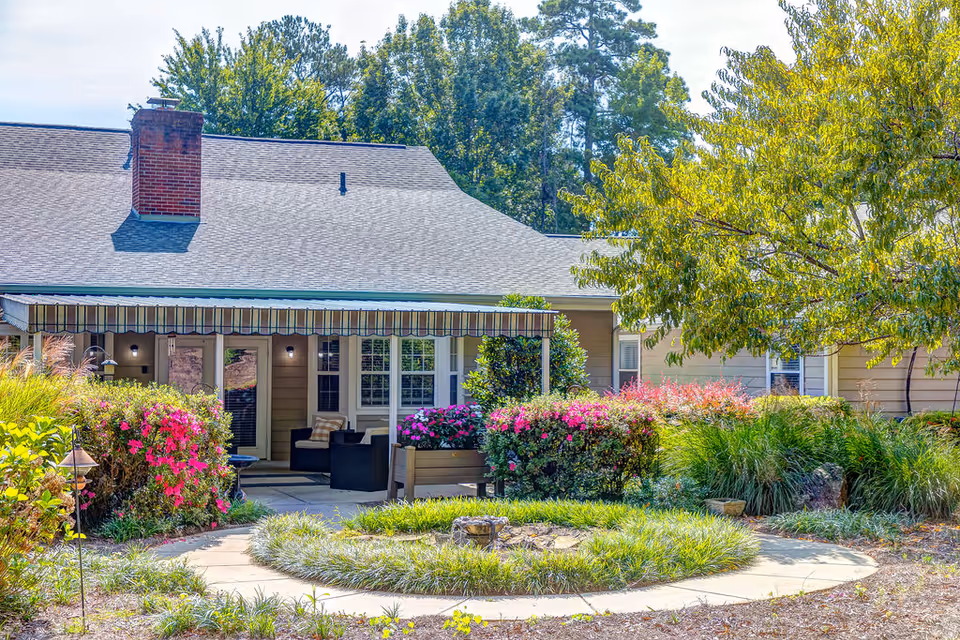 Outdoor view of a senior living facility with a covered patio area featuring outdoor seating. The patio is surrounded by vibrant flowering bushes and greenery, with a circular walkway and landscaped garden in the foreground. Trees and a clear sky are visible in the background.