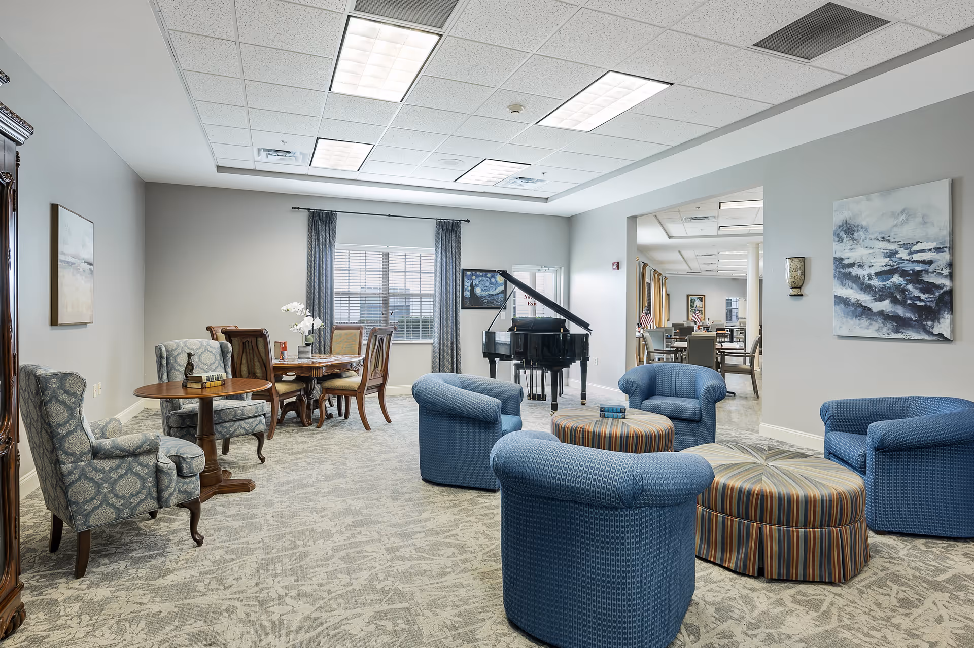 A spacious and well-lit common area in a senior living facility featuring a group of four blue upholstered chairs arranged around two round striped ottomans. In the background, there is a black grand piano near a window with gray curtains. To the left, there is a wooden dining table with chairs and a small round side table with a decorative item. The walls are painted light gray and adorned with framed artwork.
