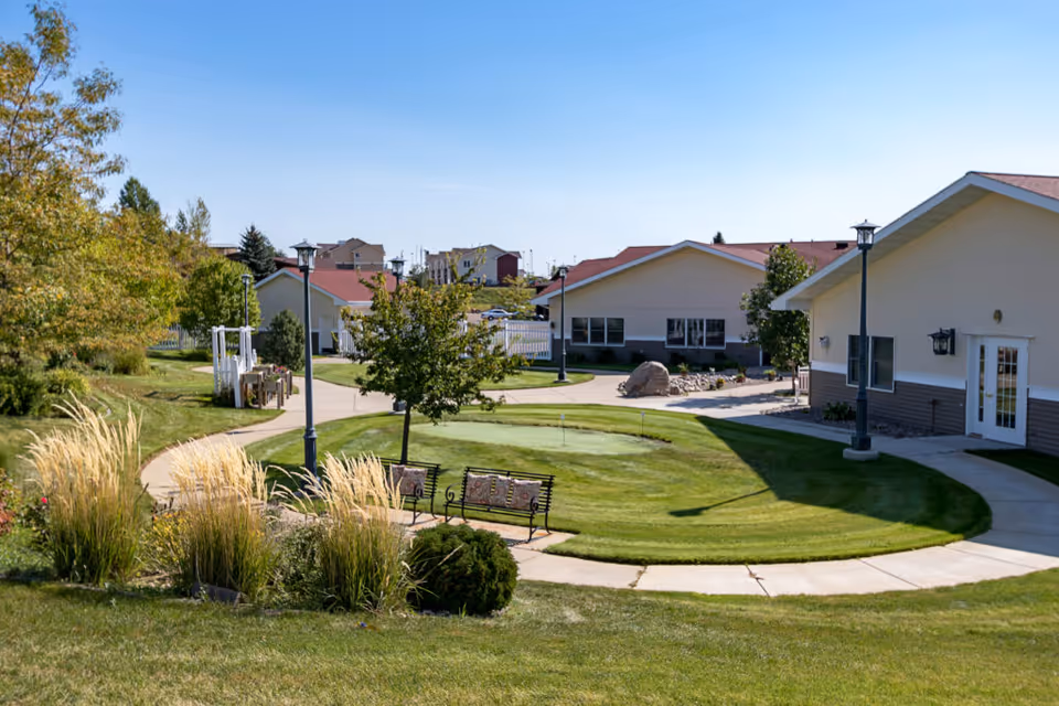 Outdoor view of a senior living facility with well-maintained green lawns, a small putting green, benches, trees, and paved walkways. Several single-story buildings with light-colored walls and red roofs are visible under a clear blue sky.
