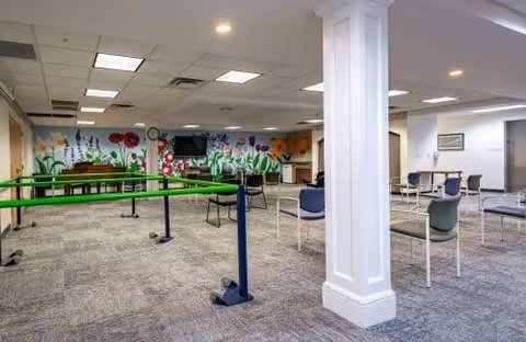 Spacious interior activity room with chairs and tables, green portable bars, and a colorful floral mural on the back wall.