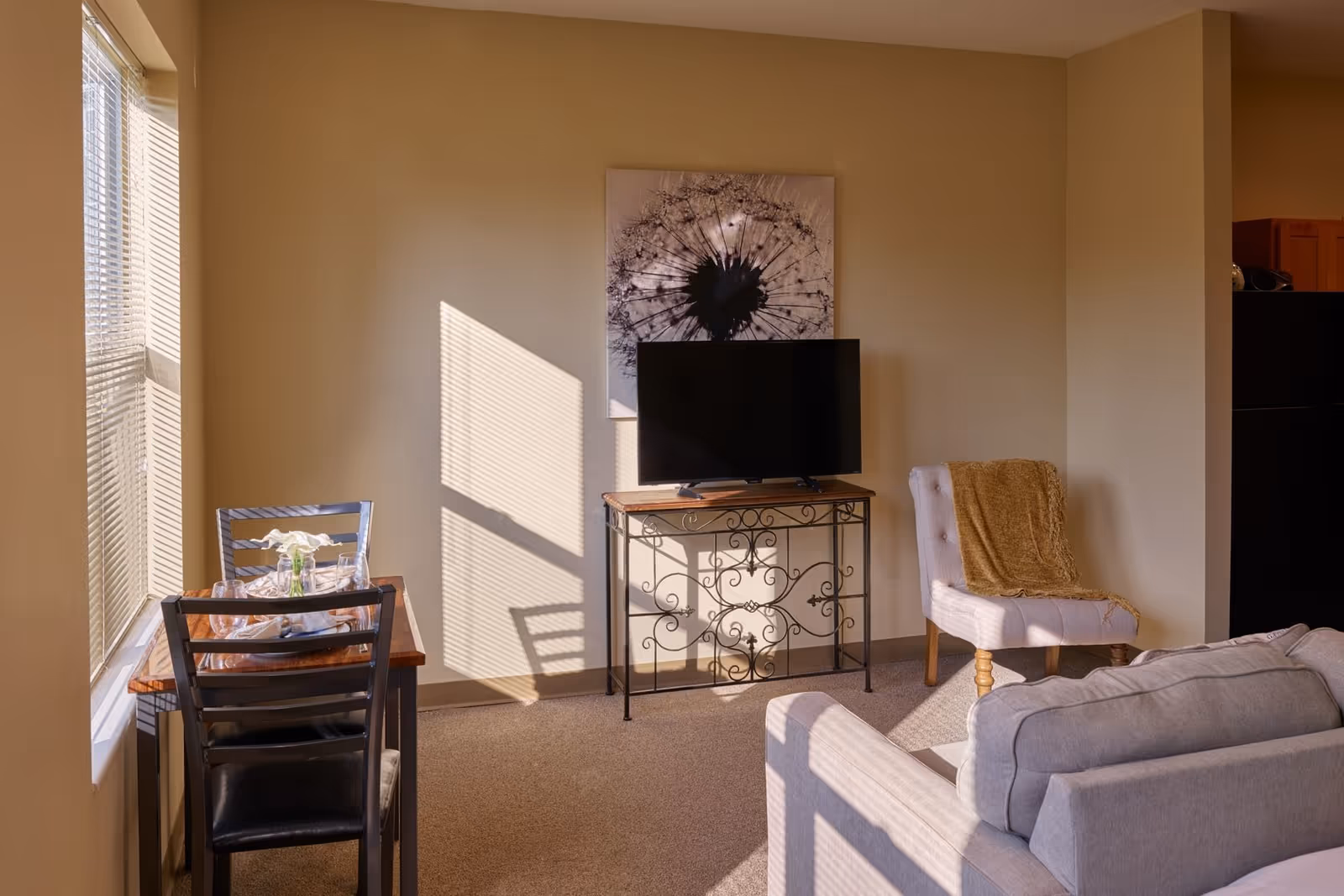 A cozy living area with a small dining table set for two near a window with blinds. A flat-screen TV sits on an ornate metal and wood stand against a beige wall with a large dandelion artwork above it. There is a cushioned chair with a brown throw blanket and part of a light gray sofa is visible in the foreground.