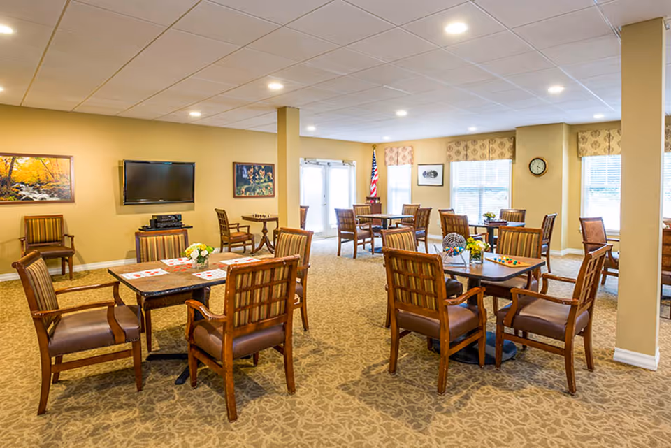 A spacious, well-lit common area with multiple tables and chairs arranged for group activities or dining. The room features beige walls, carpeted floors with a patterned design, and several windows with valances letting in natural light. There is a flat-screen TV mounted on the wall, framed artwork, an American flag near the windows, and various game items on the tables.