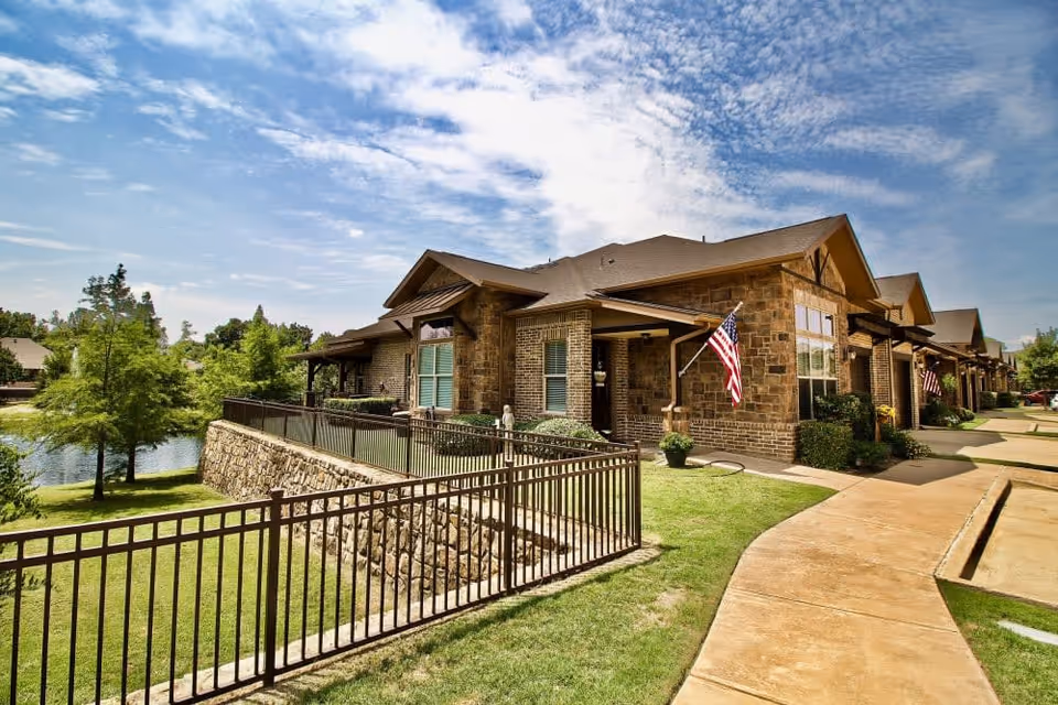 Exterior view of a single-story brick building with multiple units, each featuring a small porch with an American flag. The building is surrounded by a well-maintained lawn, a black metal fence, a stone retaining wall, and a pond with trees in the background under a partly cloudy blue sky.