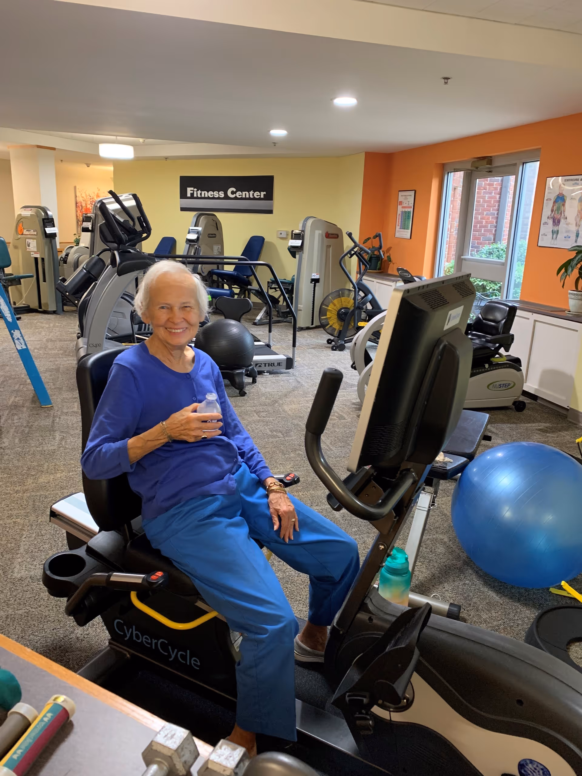 An older woman seated on a recumbent exercise bike inside a fitness center, smiling and holding a water bottle.