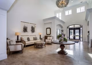 A spacious and well-lit lobby area with high ceilings and large windows. The room features a round wooden table with a decorative plant centerpiece in the middle, a seating area with a sofa, two armchairs, a round ottoman, two table lamps, and wall art. The entrance door is glass with black framing, and there is a mirror on the wall near the seating area.