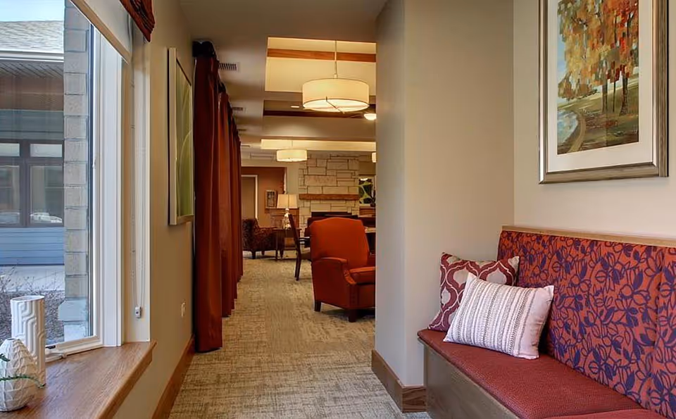 Interior common living area with a bench and pillows in the foreground, red armchairs and pendant lights down a carpeted corridor.