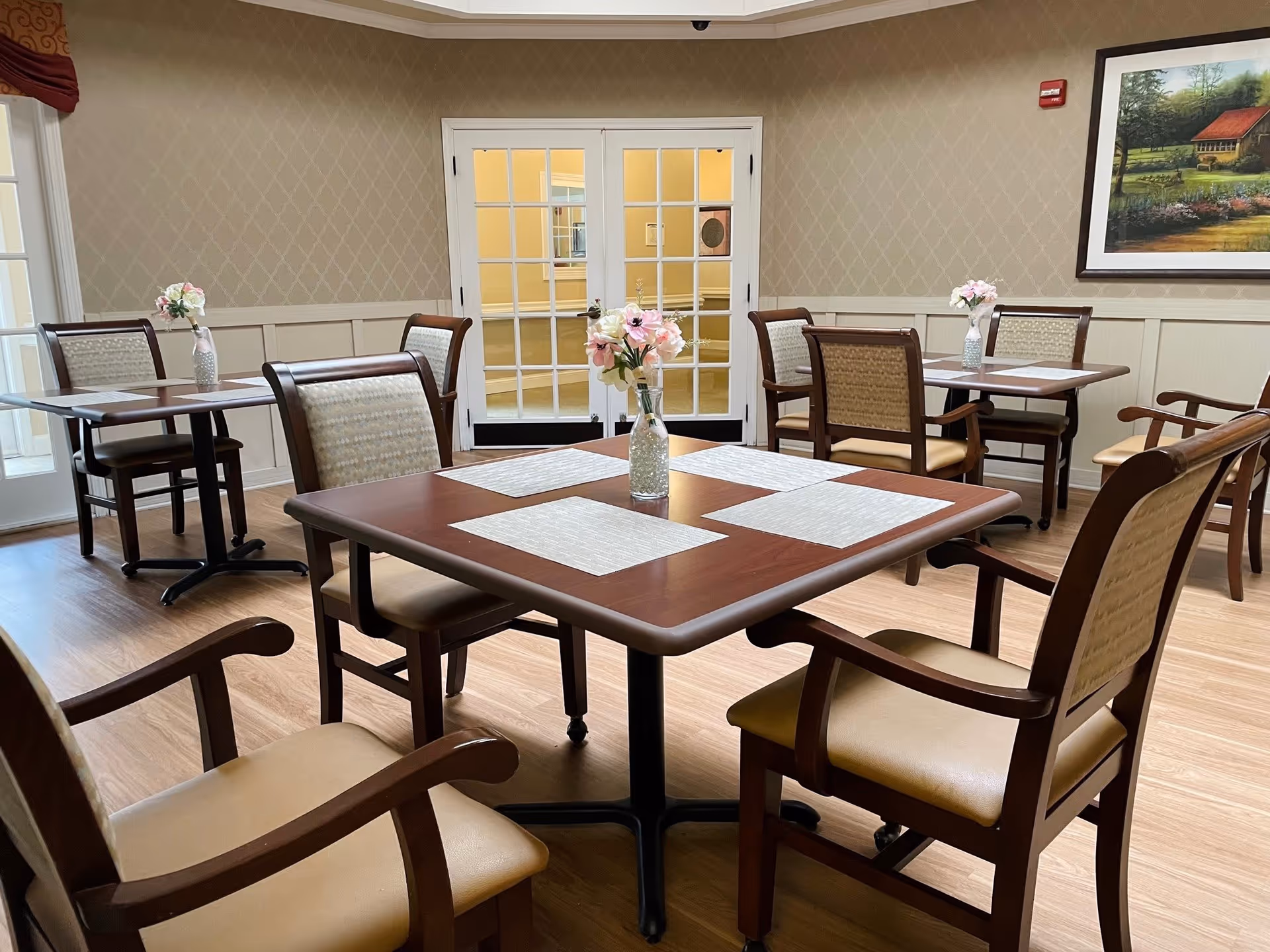 A dining room with several wooden tables and cushioned chairs arranged neatly. Each table has placemats and a small vase with pink flowers. The room has light-colored walls with a diamond pattern, wooden flooring, and a framed painting on the wall. French doors with glass panes are visible in the background.