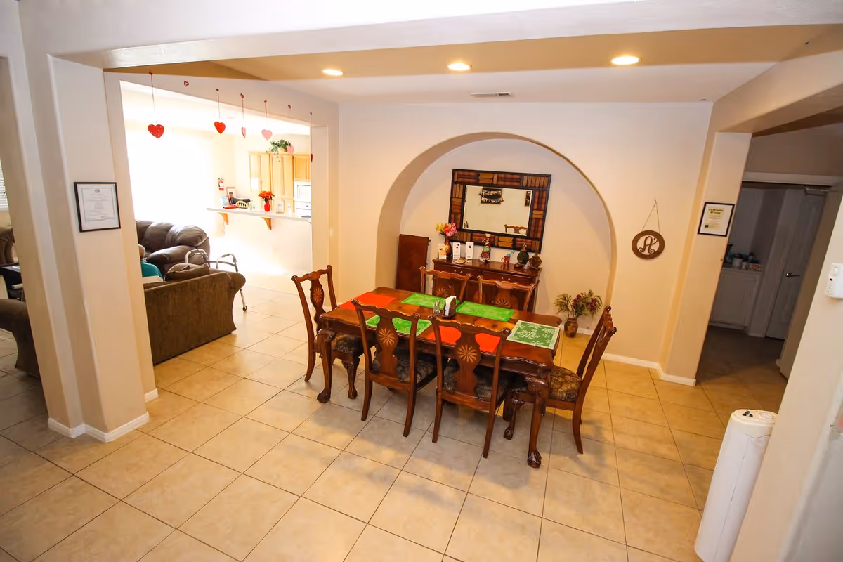 A dining area with a wooden dining table and six chairs, decorated with placemats and a centerpiece. Behind the table is a sideboard with a large mirror above it, flowers, and decorative items. The room has tiled floors and beige walls. To the left, there is a living room area with sofas and a kitchen area visible in the background. The ceiling has recessed lighting, and red heart decorations hang from the ceiling near the kitchen.