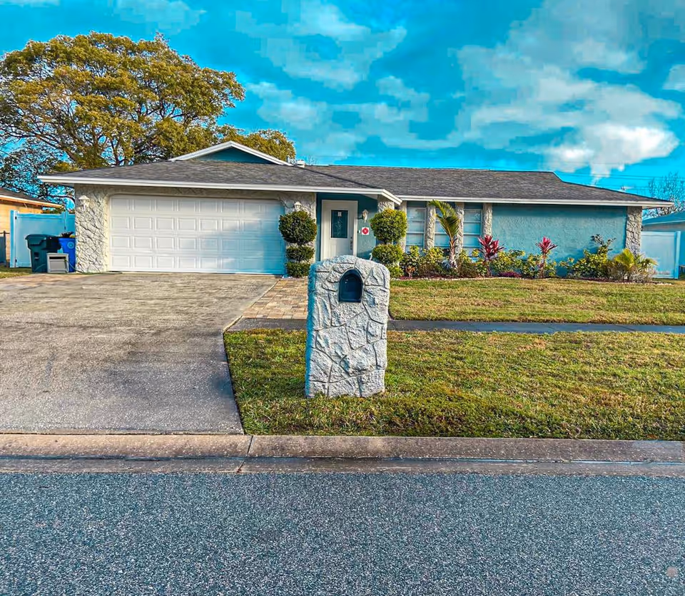 Front exterior view of a single-story house with a two-car garage, a stone mailbox, neatly trimmed bushes, and a well-maintained lawn under a partly cloudy sky.