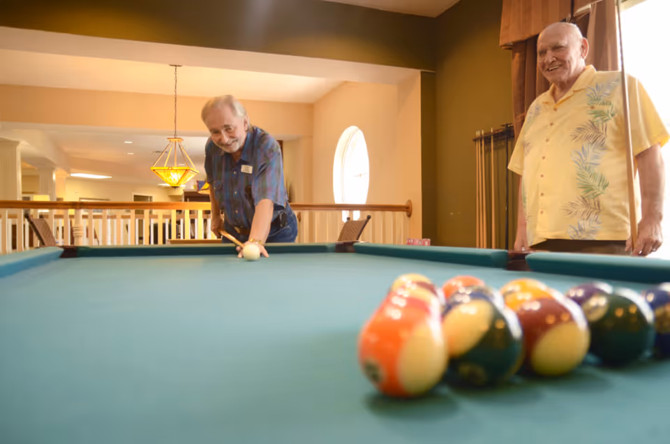 Two elderly men playing pool in a well-lit room with a green pool table. One man is lining up a shot with a cue stick while the other watches and smiles. The room has warm lighting, a hanging lamp, and a window with curtains.