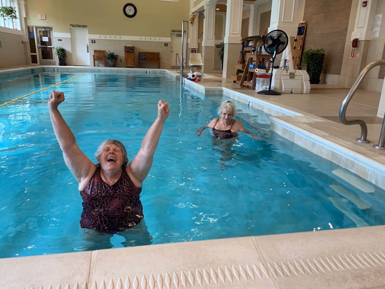 Two elderly women enjoying themselves in an indoor swimming pool at a senior living community. One woman is closer to the camera with her arms raised and smiling, while the other is further back in the pool. The pool area is well-lit with beige tiled walls and floor, benches, plants, and pool safety equipment visible.