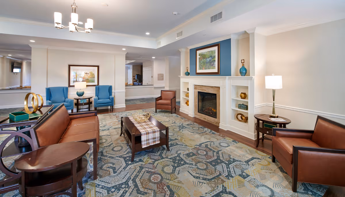 A spacious and well-lit living room area with a patterned carpet, brown leather sofas and chairs, a wooden coffee table with a decorative runner, and a white fireplace with built-in shelves displaying decorative items. The walls are light-colored with crown molding, and there are framed artworks and lamps providing additional lighting.